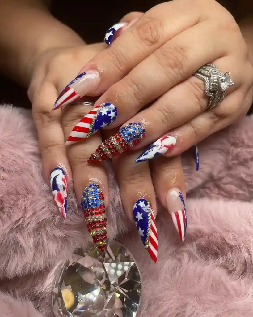 Patriotic acrylic nail designs with red, white, and blue stripes, layered glitter, and 3D embellishments like rhinestones and pearls for a dramatic look